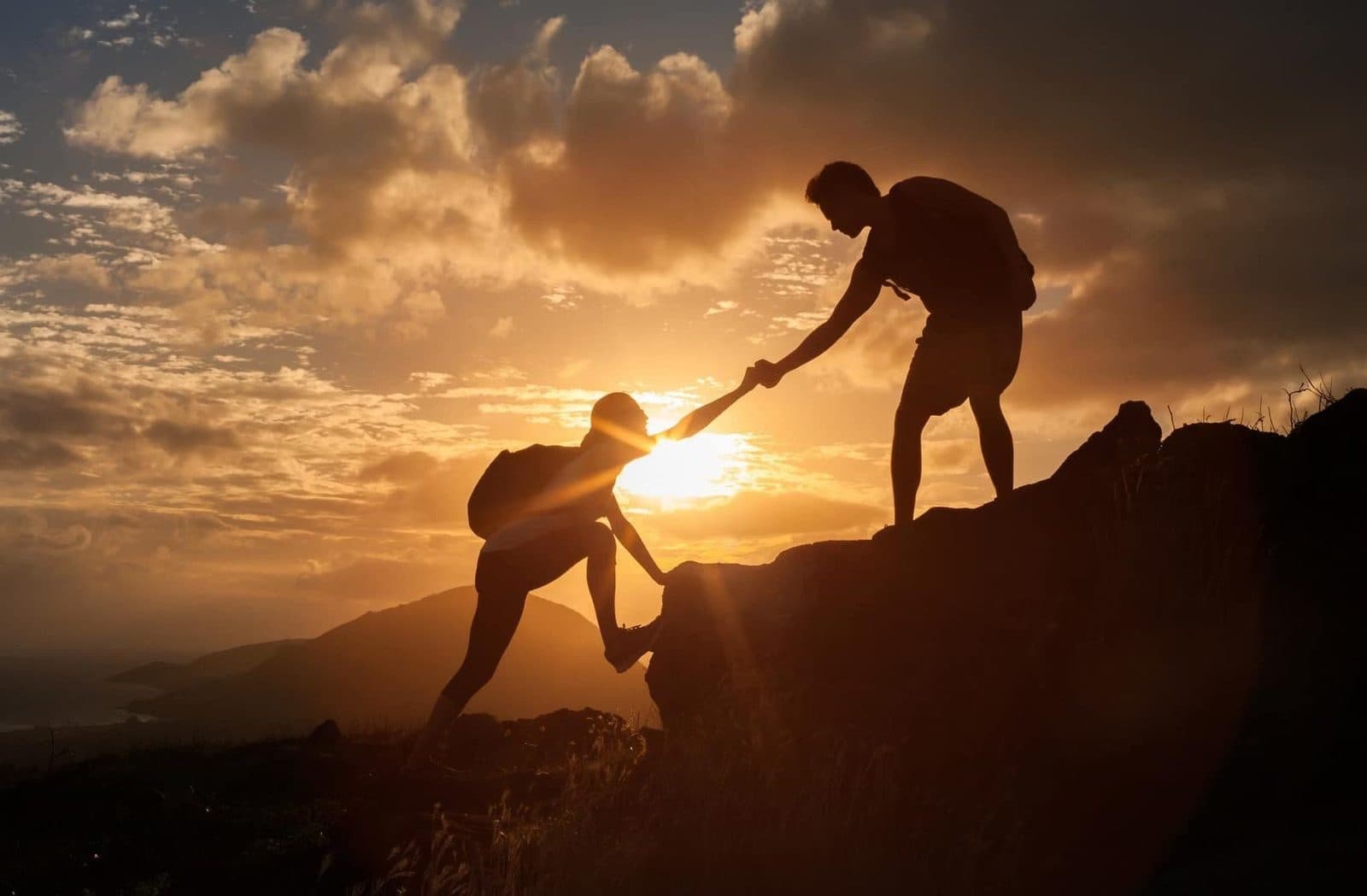 At sunset, one person reaches down to pull another up the mountainside.
